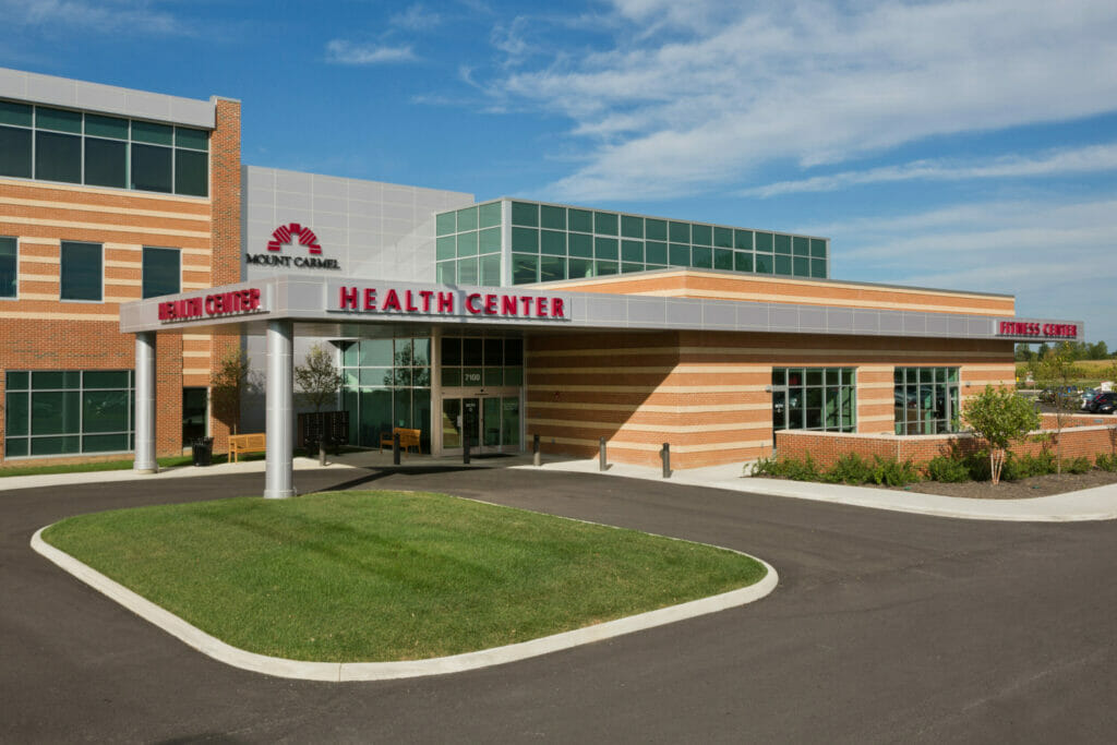 Exterior of Mount Carmel Health health center entrance with brick and tan stripped exterior with view of fitness center to the right with parking