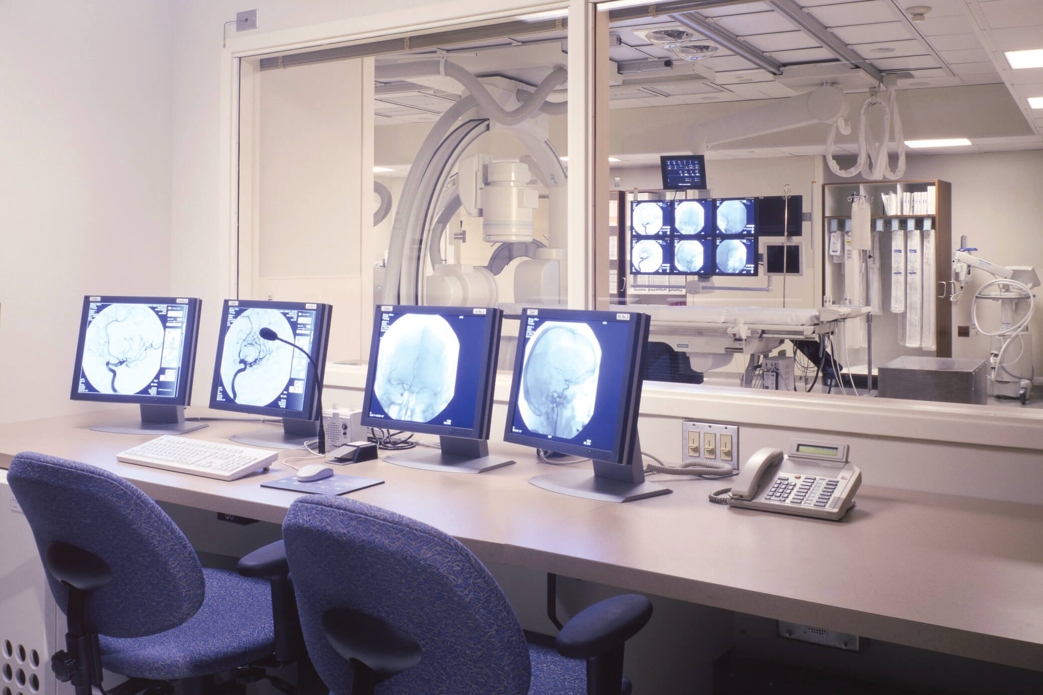 Medical exam room from the view of an attached office with multiple screens with windows looking into the room