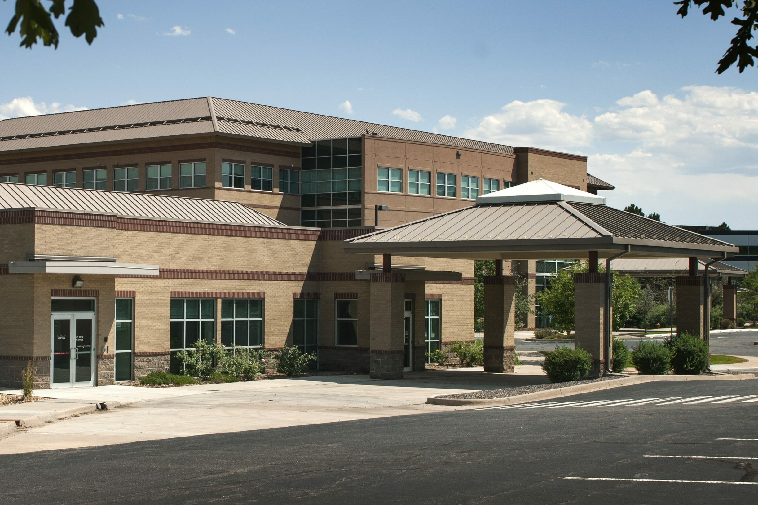 Sideview of brick building with porte-cochère with shrubs in front of the parking area and a taller, brick building in back