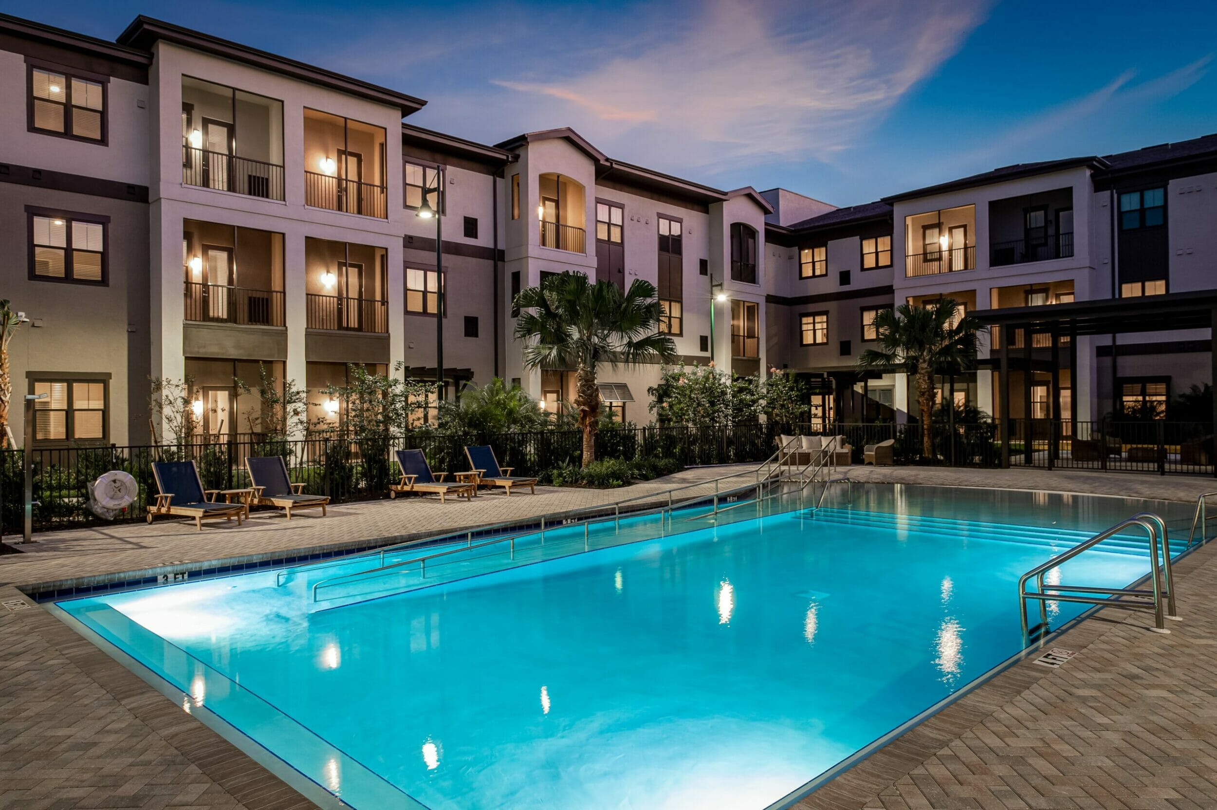 Exterior view of The Gallery at North Port pool and 3-story cream stucco community at night with lighting in pool and residences