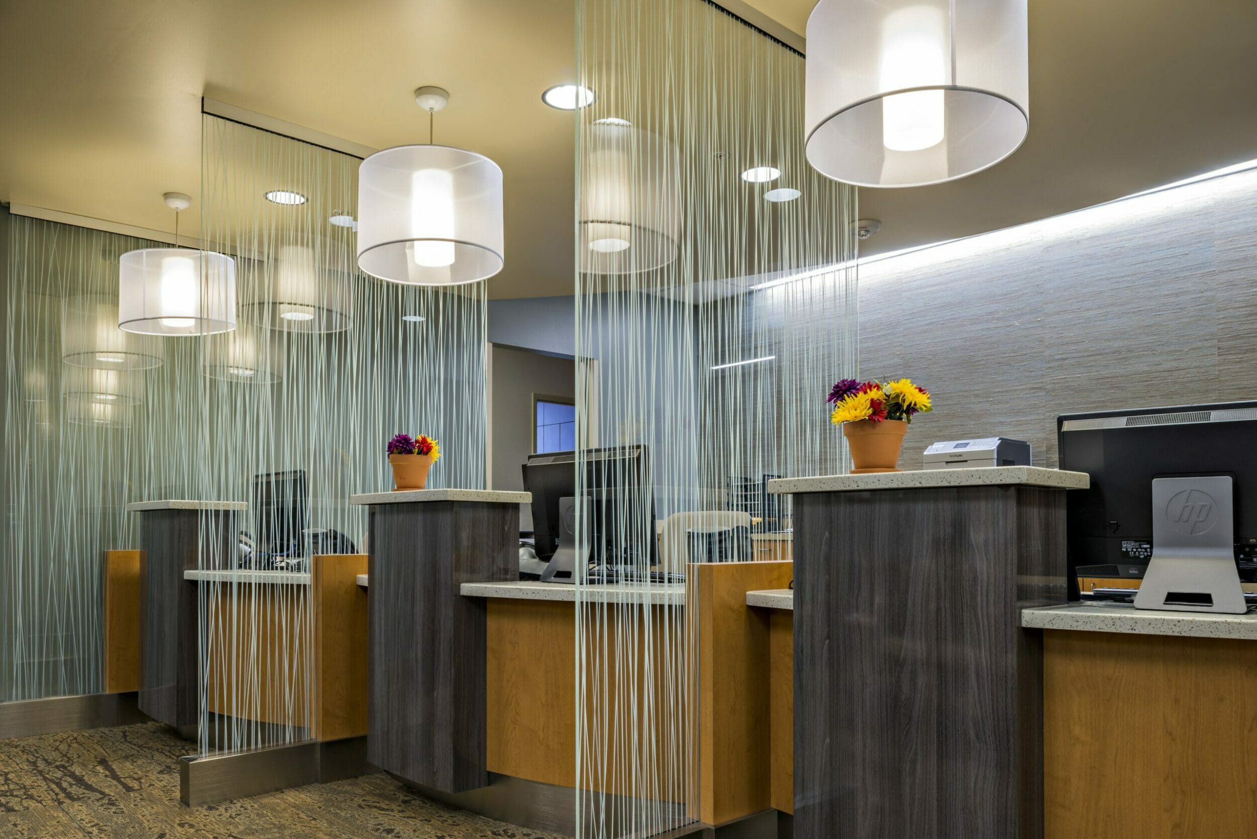 Wood front desk with three computers seperated by a glass partition wall in front of a stone wall with clear chandeliers over each computer