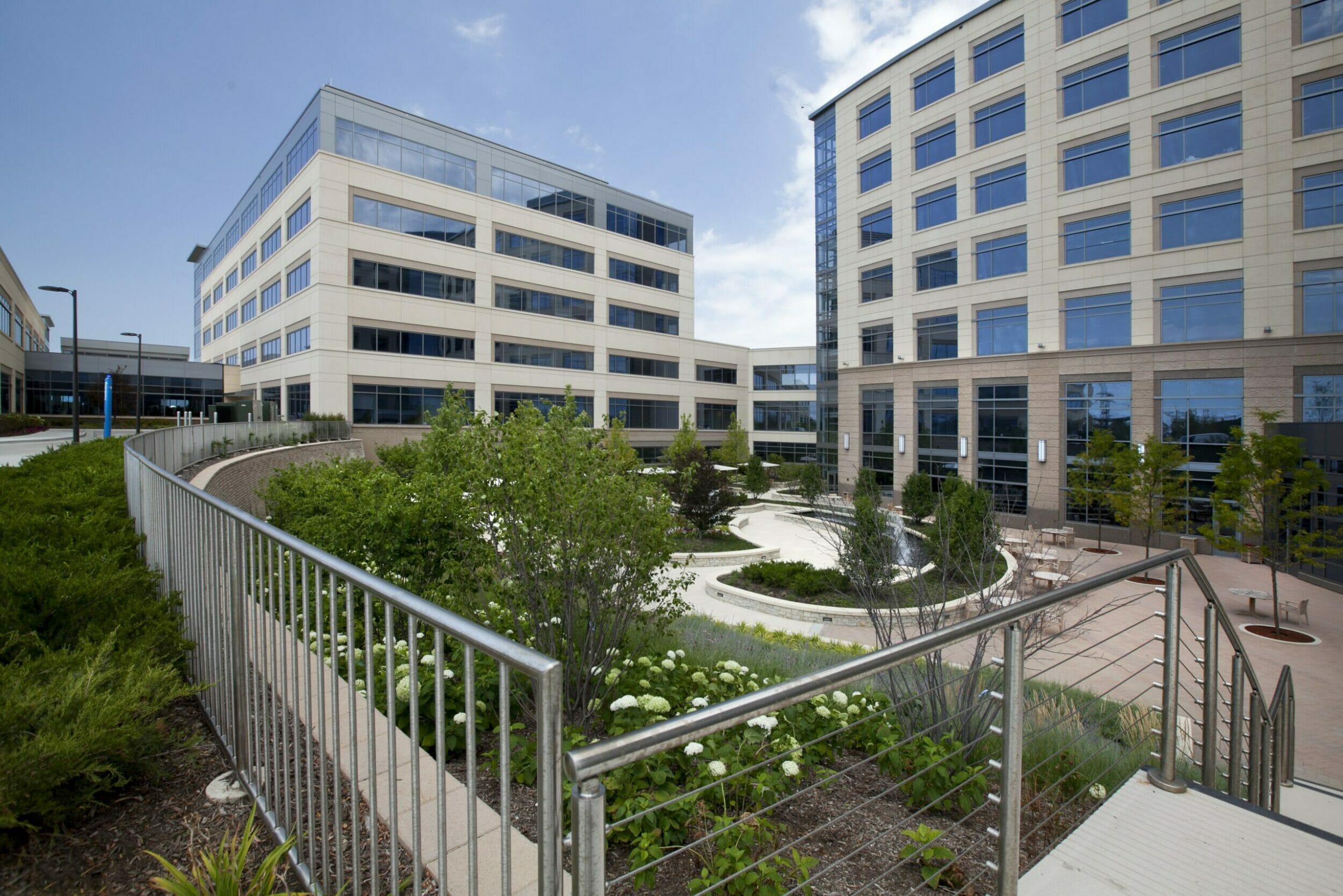 Exterior view medical office building courtyard. Lush landscaping and water fountain, tables and chairs, half shaded with umbrellas