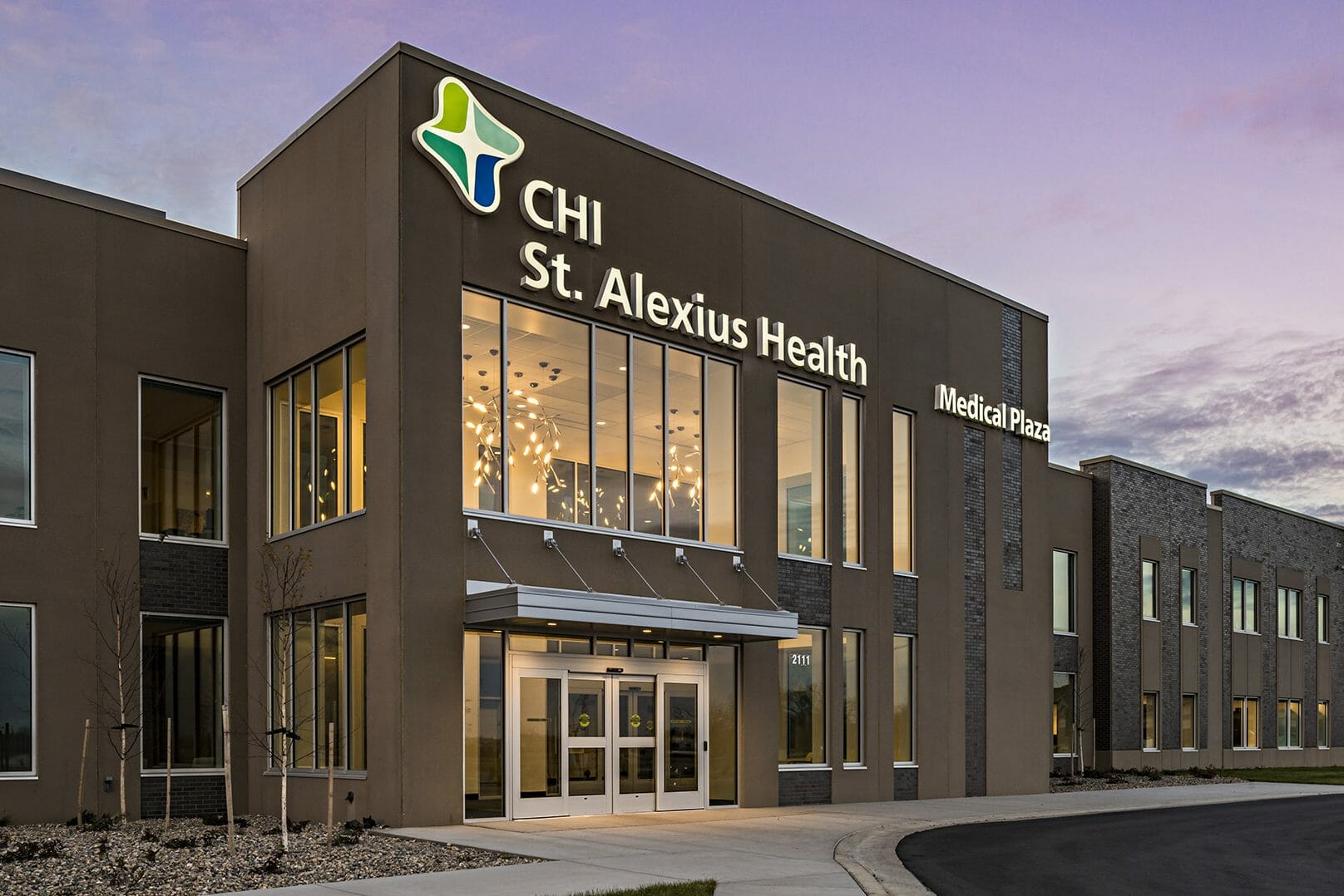 Exterior side angle of front of CHI St. Alexius Health medical plaza at night with stone and stucco finishes and a view of the parking lot