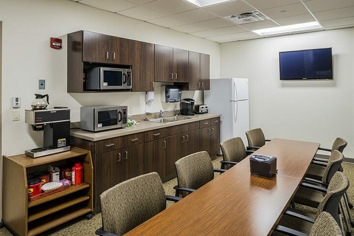 Break room inside medical office with a long, wood table with many chairs along with a mounted TV, kitchenette, and mail area