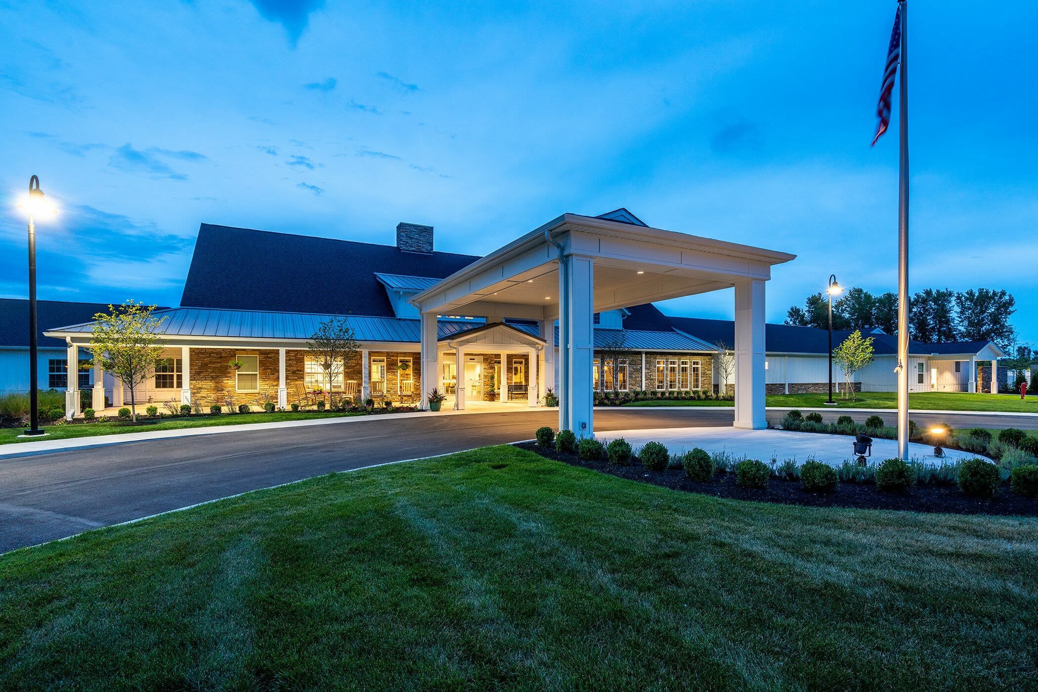 Exterior of Walnut Crossing senior living at night with american flag outside and plants lining the building that is white with stone accents